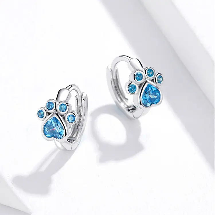 Silver hoop earrings featuring a paw design adorned with blue gemstones, including a heart-shaped center stone.