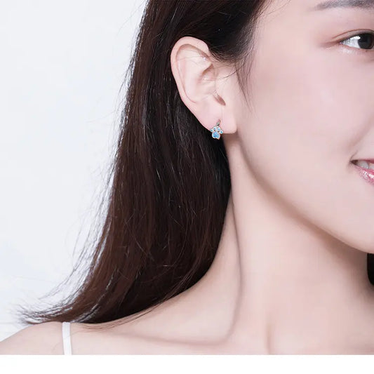 A close-up of a woman’s ear showcasing a delicate blue flower-shaped earring, complemented by her long, dark hair.